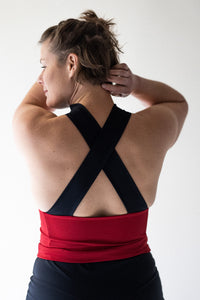 Brunette woman in studio facing away with hands in her hair wearing Davy J Sustainable Waterwear red swim top with wide black crossback straps overlapping black high waist bikini briefs rolled down to show red lining. 
