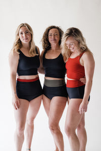 Three smiling women in studio with arms around each other all wearing Davy J Sustainable Waterwear black and red crop swim tops and black high waist bikini briefs rolled down to show red and olive green linings