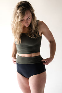 Blonde woman in studio looking down as she folds down her black high waist Davy J Sustainable Waterwear bikini briefs to show olive green lining, worn with olive green crop swim top 