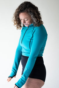 A lady with brown curly hair holds her hands out over the floor and is looking down at them with the sleeves pulled over her hands and thumbs. She is wearing a Davy J Sustainable Waterwear ocean green long sleeve swim top and black high waist bikini briefs