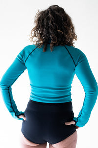 A back view of a lady with short brown curly hair and her hands on her hips, wearing a Davy J Sustainable Waterwear ocean green long sleeve swim top and black high waist bikini briefs