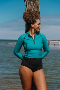 A lady with brown curly hair in a high ponytail stands against a piece of driftwood in front of the sea with her arms resting behind her back.  She's smiling and looking to the right.  She's wearing a Davy J Sustainable Waterwear ocean green long sleeve top and black high waist bikini briefs 