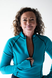 A lady with short brown curly hair looks directly into the camera smiling.  She wears an ocean green Davy J Sustainable Waterwear long sleeve swim top and is holding the zip on the front pulled down.  The sleeve is long and has visible thumb holes.