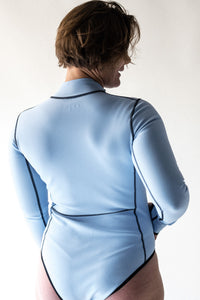 A back view of a lady with short brown hair wearing a Davy J Sustainable Waterwear powder blue long sleeve swimsuit with black stitching detail