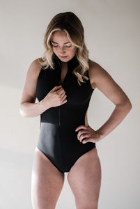 Blonde woman in studio looking down as she touches zip on the front of her black Davy J Sustainable Waterwear swimsuit with cut out back 