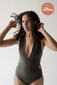 Woman in studio with hands in her long curly brown hair, wearing an olive green Davy J Sustainable Waterwear cutout swimsuit with plunge neckline, and hoop earrings