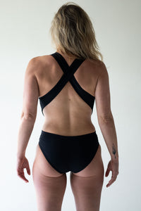 Blonde woman in studio facing away with hair over one shoulder, wearing black Davy J Sustainable Waterwear cutout swimsuit with cross back straps 