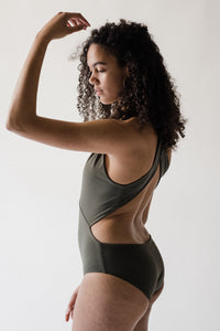 Woman with long curly brown hair in studio facing side on with one arm raised wearing an olive green Davy J Sustainable Waterwear cutout swimsuit with plunge neckline and wide cross back straps, and hoop earrings