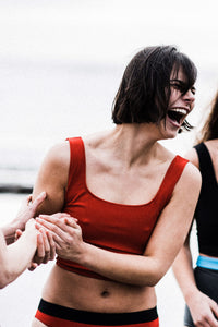 A woman laughing with short black bobbed hair holds hands with another woman.  She is wearing a red Davy J Sustainable Waterwear cropped swim top with square neckline and shoulder straps and a pair of low rise red bikini briefs with black waistband
