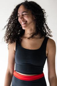 A lady with black curly hair smiling, wearing a Davy J Sustainable Waterwear black cropped swim top with shoulder straps and black high waist briefs with waist rolled down and showing red lining