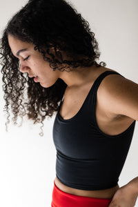 Side view of a lady with long black curly hair, looking down at the floor and hand on hip, wearing a Davy J Sustainable Waterwear black cropped swim top with square neckline and shoulder straps. 