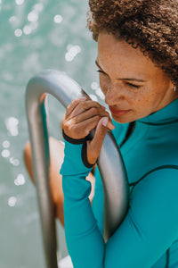 A woman with brown curly hair sat on steps going into the sea, wearing a Davy J Sustainable Waterwear ocean green long sleeve swimsuit.  She is holding on to the handrail of the steps, showing the thumb detail of the sleeve