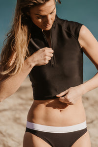 A slim lady with blonde hair standing in front of a rock pulls the zip up on a Davy J Sustainable Waterwear black short sleeve swim top.  She also wears a pair of low rise black bikini briefs with white waistband