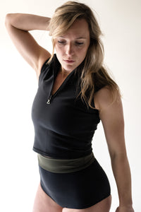 A blonde lady facing down with her hand on her head, wearing a Davy J Sustainable Waterwear black short sleeve swim top with half zip and a pair of black high waist bikini briefs with the waist folded down to show the olive lining